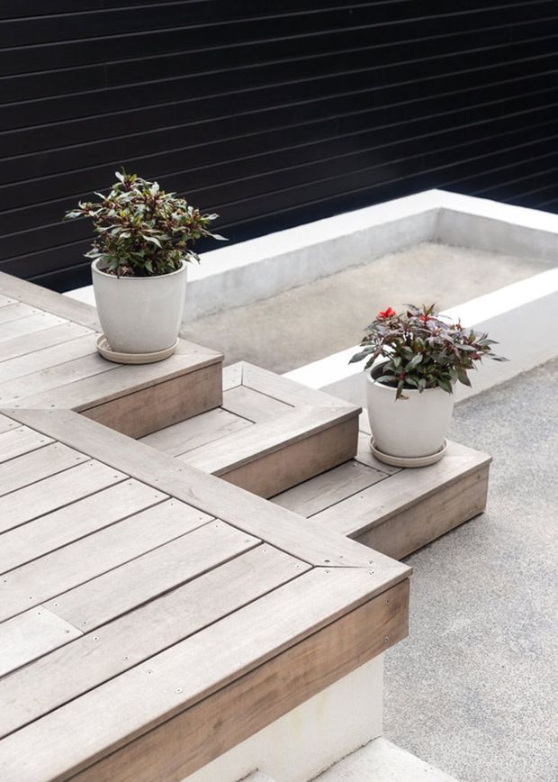 A contemporary outdoor terrace featuring stepped timber decking platforms, integrated raised seating or planting tiers, white rendered concrete edges, and potted shrubs. A dark horizontal-clad wall forms the backdrop, creating a clean, geometric courtyard-like setting.
