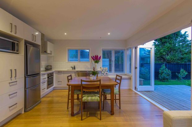 Bright open-plan kitchen and dining area with white built-in cabinetry, stainless-steel appliances, timber flooring, a compact timber dining table and chairs, and extensive glazing opening onto a timber deck and landscaped garden.