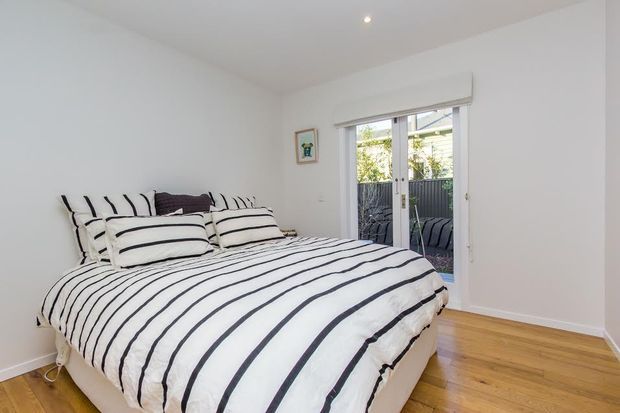 Bright, compact bedroom with a neatly made bed dressed in white bedding with bold navy or dark grey horizontal stripes. The room has white walls, recessed ceiling lighting, timber flooring, and glazed French doors opening to a small exterior area or balcony. Minimal furnishings create a clean, relaxed look.