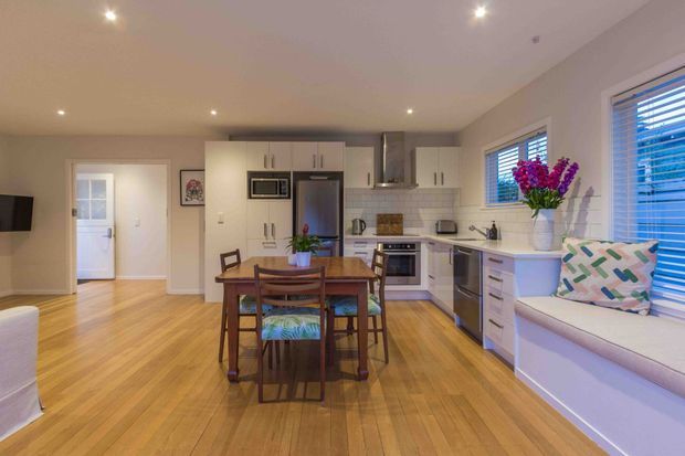 Open-plan kitchen and dining area with light cabinetry, stainless-steel appliances, timber flooring, a wooden dining table and chairs, and a built-in window seat with cushions. Bright recessed lighting and large windows with blinds create a warm, inviting interior.