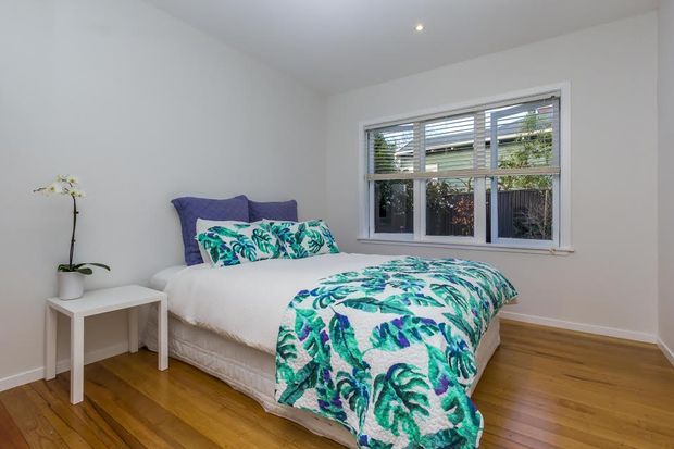 Bright, compact bedroom with white walls and ceiling, a double bed dressed in white bedding and a vivid teal, green and purple botanical duvet, a small white bedside table with an orchid, and warm timber flooring. A wide window with horizontal blinds provides natural light and views of greenery outside.