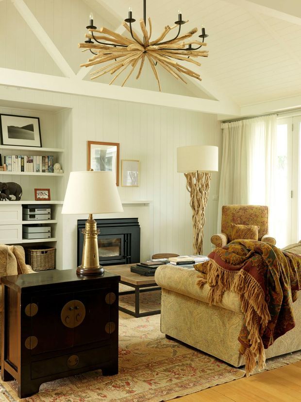 Warm, inviting living room with white vertical wall panelling, built-in shelving and cabinetry, a black fireplace, exposed angled ceiling beams, and bright glazing with sheer curtains. The room is furnished with a patterned sofa, upholstered armchair, coffee table, dark timber side cabinet, ornate lamps, and a striking branch-like chandelier. A layered vintage-style rug and warm timber flooring add texture and character.
