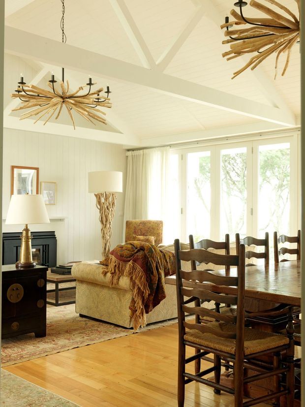 A warm, light-filled open-plan living and dining space with a white painted timber-paneled interior, exposed vaulted ceiling beams, and large glazed doors. The room features a rustic timber dining table and ladder-back chairs, upholstered armchairs, a dark fireplace surround, timber flooring, vintage-style rugs, and distinctive branch-like statement chandeliers.