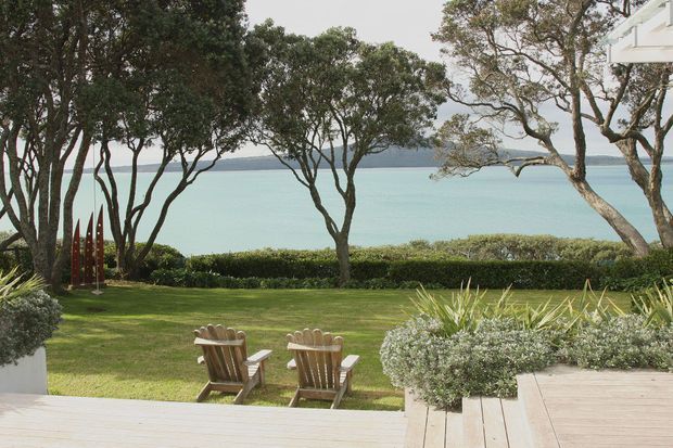 A landscaped coastal outdoor area viewed from a timber deck, with two wooden Adirondack-style chairs facing a calm turquoise bay. Mature trees, clipped hedges, lawn, sculptural planting, and a white building edge frame the waterfront outlook.