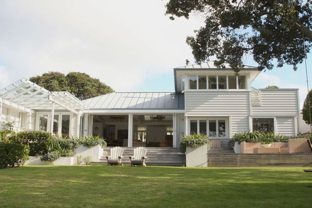 A broad exterior view of a refined coastal-style residence with white horizontal weatherboard cladding, pale standing-seam metal roofs, extensive glazed openings, and a glazed conservatory or veranda to the left. The central covered outdoor living area opens onto steps and a manicured lawn, with raised planting beds and mature trees framing the house.
