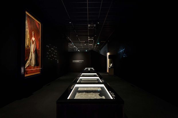 A dark, contemporary museum or gallery exhibition interior with black walls and ceiling, dramatic focused lighting, a large framed artwork on the left wall, and illuminated horizontal display cases arranged along the central walkway.