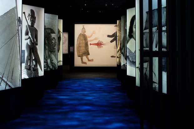 A dark immersive exhibition gallery or museum installation with illuminated vertical art panels arranged along both sides of a passage. The central projection depicts stylised figures and a red hand motif, while blue rippling light covers the floor and black structural framing encloses the display.