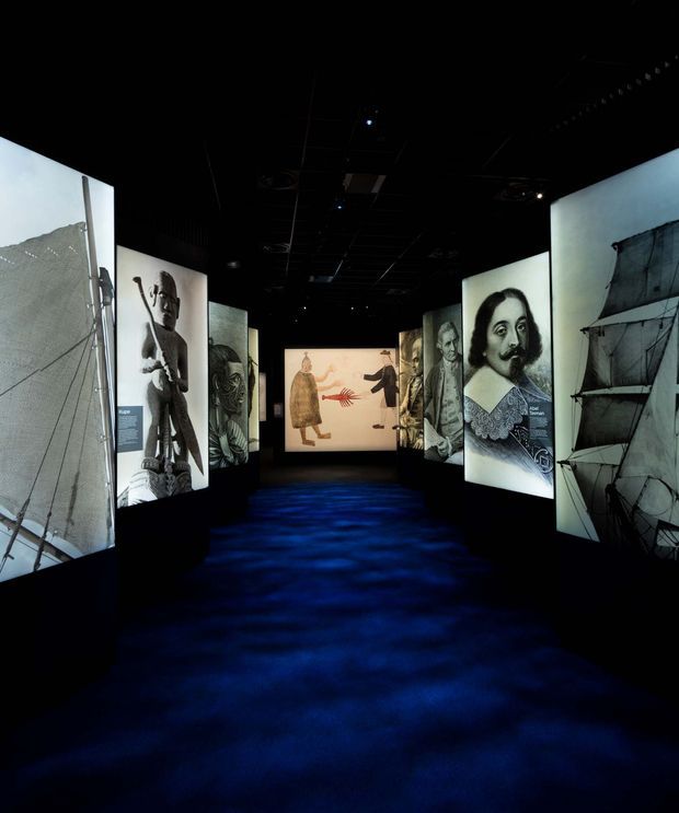 A dark immersive exhibition gallery with large illuminated panels displaying historical portraits, illustrations, diagrams, and projected imagery. The space has a black ceiling and walls, a deep blue carpeted floor, and a dramatic museum-like presentation with freestanding display screens arranged along a central walkway.
