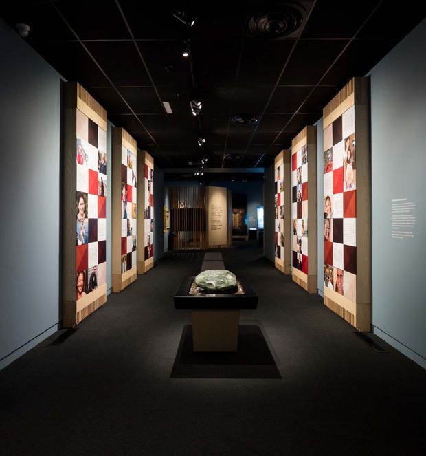 A dark, immersive exhibition or gallery corridor with black ceiling, dark carpeted flooring, and illuminated freestanding display walls on both sides. The displays combine portrait photography, red-and-white graphic panels, timber-toned framing, and small blocks of text. A central pedestal presents a sculptural object, while track lighting and recessed services illuminate the passage.