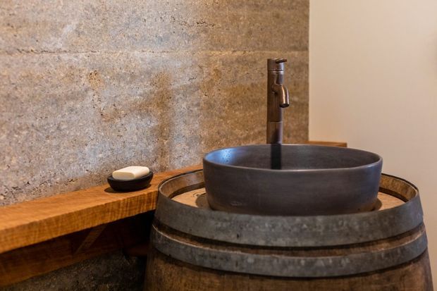 Close-up of a rustic bathroom vanity featuring a dark vessel basin mounted on a repurposed wooden barrel, with a bronze-toned wall-mounted tap and a thick live-edge timber shelf holding a soap dish. The backdrop combines textured stone or concrete-look wall surfacing with warm natural wood and dark metal details.