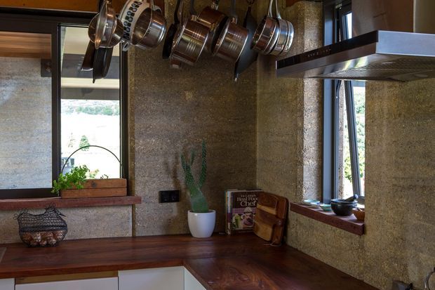 Compact rustic kitchen corner with dark timber benchtops, pale lower cabinetry, textured earthen-toned wall finishes, deep timber window sills, and black-framed windows. Copper and stainless-steel cookware hangs above the work area, with a stainless rangehood and small decorative items on the counters.