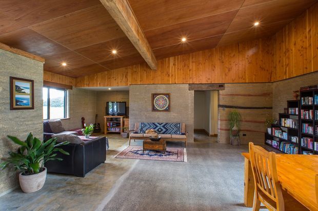 Rustic open-plan living and dining room with a concrete floor, textured earth-toned walls, extensive warm timber wall and ceiling lining, and a prominent exposed timber beam. The space includes a lounge seating area, patterned sofa, television, bookshelves, artwork, indoor plants, and a timber dining table and chairs. Recessed lighting creates a warm, inviting atmosphere.