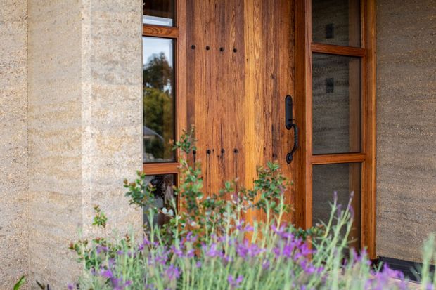 Close-up exterior view of a rustic timber entry door with dark metal hardware, set within warm timber-framed glazing and textured earthen or concrete walls. Purple flowering plants partially obscure the lower foreground, creating an earthy, natural approach.