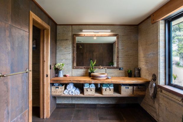A warm, rustic bathroom vanity area with a long timber countertop, open woven storage baskets, folded towels, a vessel basin, wall-mounted mirror, and exposed textured stone or concrete walls. Dark tiled flooring and timber-framed openings reinforce the natural, earthy character. A large window provides views to greenery outside.