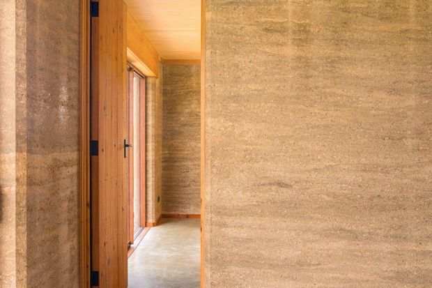 A narrow interior passage framed by thick, textured earthen walls, with a warm timber pivot or entry door and timber-lined ceiling. Natural light enters through a slim glazed opening, highlighting the raw, tactile wall surfaces and polished concrete-like floor. The view appears to be from within an entry or hallway.