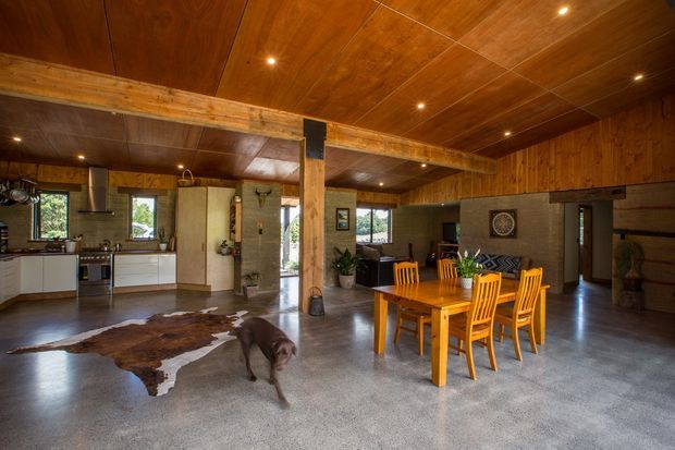 Open-plan rustic living and dining area with an integrated kitchen, featuring extensive warm timber wall and ceiling lining, exposed timber beams and posts, polished concrete flooring, a wooden dining table with chairs, and a lounge area beyond. Large windows bring in natural light and views of greenery.