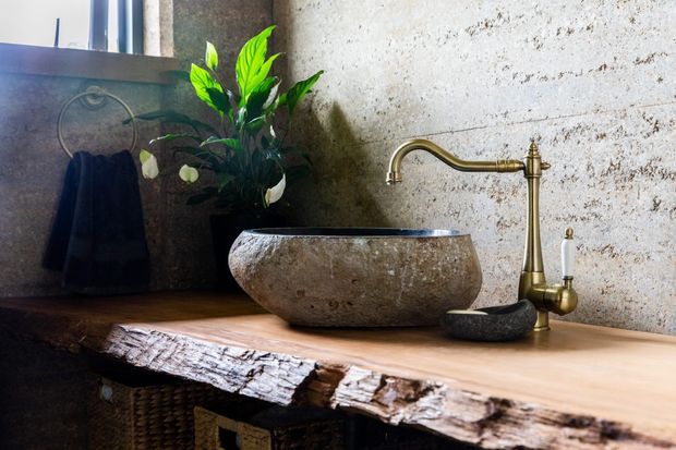 Rustic bathroom vanity featuring a rough-hewn natural stone vessel basin on a thick live-edge timber countertop, paired with an antique-style brass faucet. A textured stone wall, dark towel, woven storage, and potted greenery create an organic, earthy setting.
