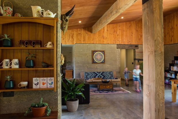 Rustic open-plan living area with exposed timber wall and ceiling lining, substantial timber beams and posts, rough grey masonry walls, polished concrete flooring, built-in timber shelving displaying ceramics, and a lounge seating area around a low wooden coffee table. A hallway or adjacent space is visible beyond the living room.