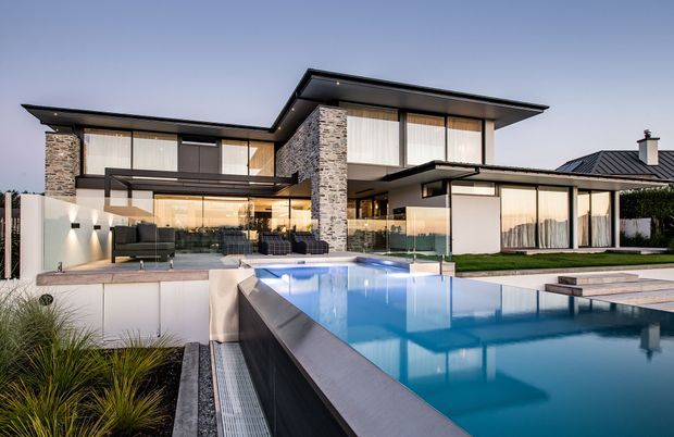 Contemporary multi-level luxury residence with extensive floor-to-ceiling glazing, dark metal rooflines and frames, natural stone-clad feature walls, and a large illuminated swimming pool in the foreground. The outdoor entertaining area includes lounge seating, glass pool fencing, landscaped planting, and broad paved terraces.