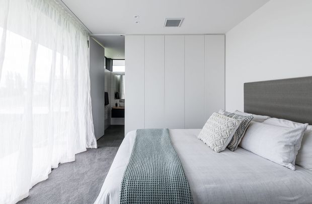 Bright contemporary bedroom with a large upholstered bed, white bedding, textured grey-blue throw, sheer full-height curtains, grey carpet, and extensive white built-in wardrobes. A compact ensuite or dressing area is visible through the opening at the rear.