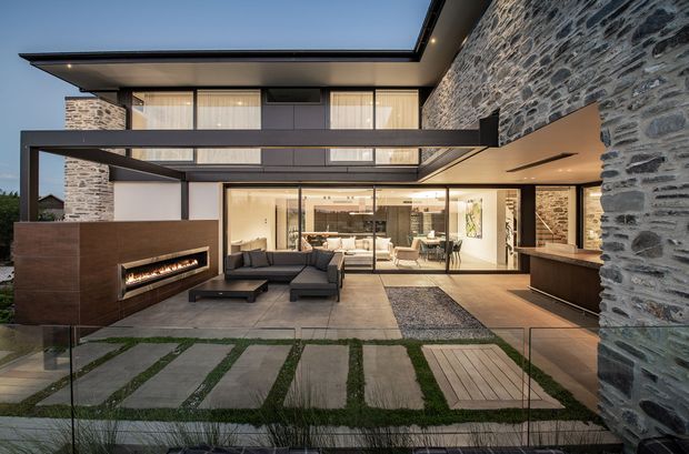 Contemporary multi-level residence viewed from a landscaped patio, featuring extensive rough natural-stone walls, dark framed floor-to-ceiling glazing, broad roof overhangs, and a covered outdoor living area. A built-in linear fireplace and seating occupy the foreground, while a warmly lit living and dining space is visible through the glazing. The patio combines large concrete pavers, narrow grass strips, timber decking, and glass balustrades.