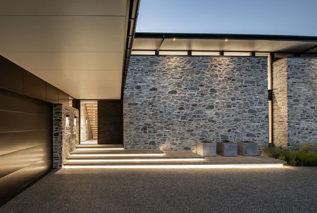 Contemporary architectural entry courtyard with a covered walkway, broad illuminated steps, dark metal wall panels, and prominent rough-stone masonry walls. Minimal planters and integrated linear lighting create a refined, dramatic approach to the building.