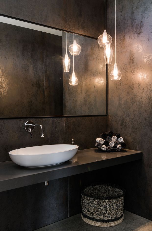 A dramatic contemporary bathroom featuring a white vessel basin on a dark floating vanity, a wall-mounted chrome tap, large mirror, dark textured wall finishes, and multiple warm-glowing glass pendant lights. A woven basket and rolled towels add texture beneath and beside the vanity.