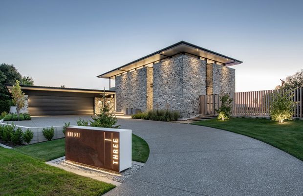 Contemporary single-storey residential facade at dusk, featuring a prominent textured grey stone-clad entry volume, broad dark roof overhangs with integrated lighting, dark garage doors, timber privacy screening, landscaped lawns and a curved concrete driveway. A sculptural illuminated address sign sits in the foreground.