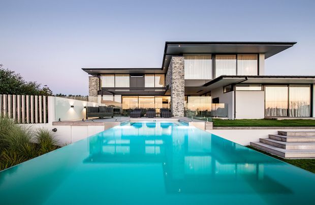 Contemporary multi-level residence viewed from a large turquoise swimming pool. The home features strong horizontal roof planes, extensive glazing, white rendered walls, textured stacked-stone cladding, dark window frames, and landscaped terraces with steps leading to the pool.