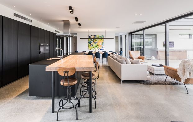 Open-plan contemporary kitchen and living area with a long timber-topped island, dark built-in cabinetry, black metal bar stools, polished concrete flooring, a neutral sectional sofa, and extensive floor-to-ceiling glazing. A dining area and colourful artwork are visible beyond the kitchen, with bright natural light throughout.