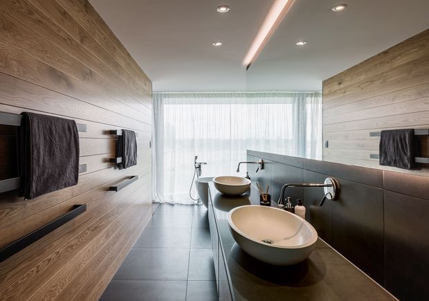 Contemporary bathroom with a long double vanity, two round vessel basins, wall-mounted taps, dark stone-look benchtop, and extensive horizontal timber wall panelling. A full-height mirrored wall reflects the vanity and fittings, while sheer curtains provide soft daylight at the far end. Recessed ceiling lights and a linear concealed light add a refined, modern feel.