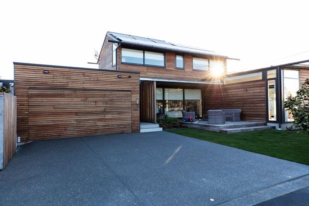 Exterior view of a contemporary two-storey residence with extensive horizontal timber cladding, a pitched metal roof, large upper-level windows, and a covered ground-floor outdoor living area. The foreground includes a broad driveway, attached timber-clad garage, paved patio, and lawn.