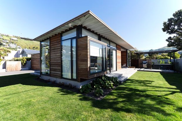 Contemporary single-storey pavilion-style house with a prominent asymmetrical pitched roof, extensive floor-to-ceiling glazing, and horizontal warm timber cladding. The building opens onto a manicured lawn and paved terrace, with a covered outdoor seating area to the side. A sunny suburban hillside setting is visible in the background.