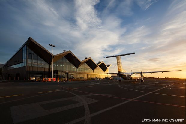 Exterior architectural photograph of a modern airport terminal at sunset, featuring a dramatic repeating zigzag roofline, extensive floor-to-ceiling glazing, dark facade cladding, and an aircraft parked on the broad tarmac in front. The image has strong warm evening light and a dramatic sky.