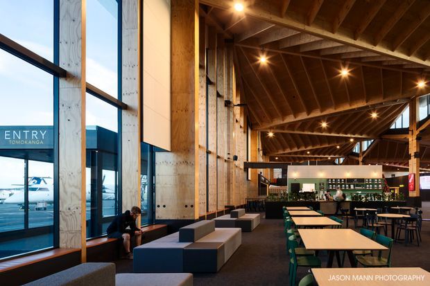 A spacious contemporary hospitality or commercial interior with a dramatic exposed timber roof structure, tall floor-to-ceiling glazing, and a large open lounge and dining area. The space includes built-in and freestanding seating, long communal tables, café-style dining furniture, and a bar or service counter at the rear. A glazed entrance marked “ENTRY TOMOKANGA” is visible on the left.