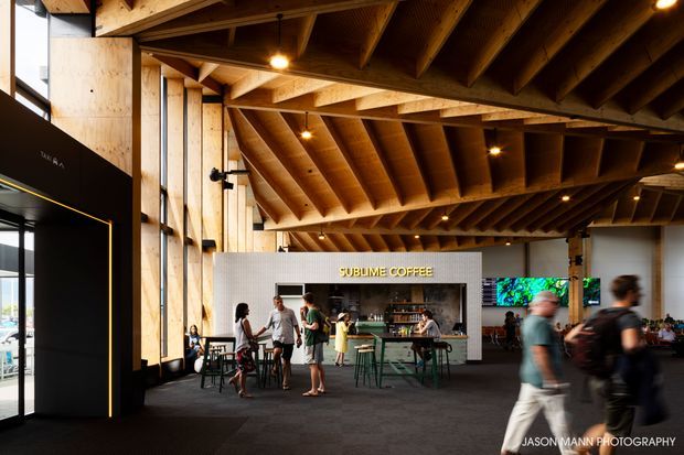 Interior view of a contemporary commercial coffee venue with a prominent exposed timber beam-and-rafter ceiling, timber structural columns, dark carpeted flooring, and a central coffee bar. Casual tables and stools occupy the open space, with large glazing at the left and illuminated screens toward the rear. People are present but secondary to the architecture and fit-out.