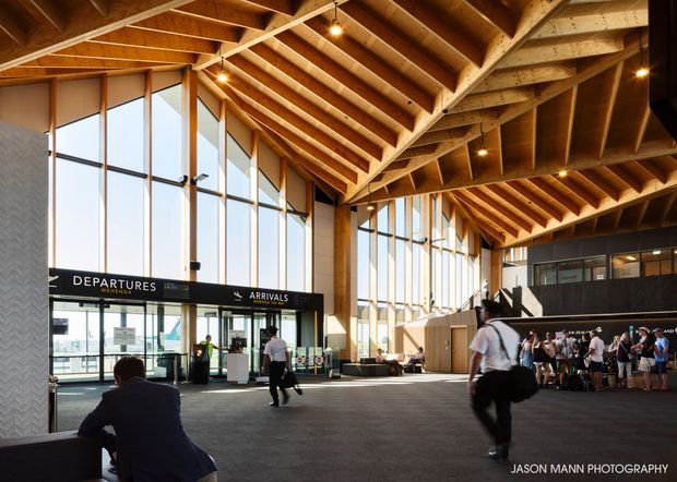 A large airport terminal interior with a spacious departure and arrivals concourse, tall floor-to-ceiling glazing, exposed timber roof trusses and beams, dark carpeted flooring, and warm pendant and spot lighting. Passengers and airport signage are visible, with a contemporary timber-framed public architecture design.