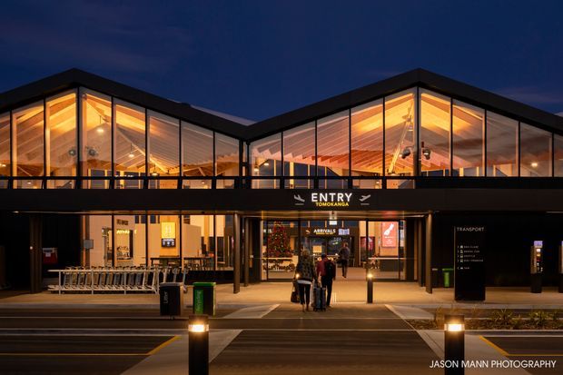 Night-time exterior view of the Entry terminal at Tongariro, showing a contemporary low-rise building with expansive floor-to-ceiling glazed façades, warm interior lighting, dark cladding, and a covered pedestrian entrance. Airport-style signage, luggage trolleys, and transport facilities are visible at the frontage.