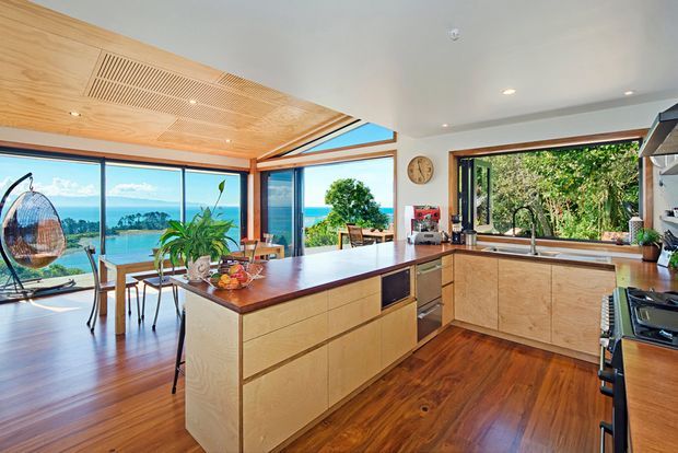 A bright open-plan kitchen with a large timber island, pale wood cabinetry, dark timber benchtops, and warm timber flooring. Full-height corner glazing opens to expansive coastal and water views, while a slatted timber ceiling feature and indoor plants add warmth and texture.