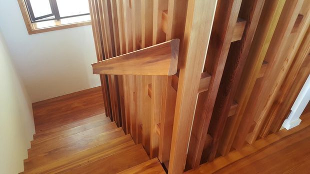 Interior view looking down a timber staircase with richly stained wooden treads, balustrade, handrail, and vertical timber slat screening. A small window provides natural light, and matching timber flooring continues at the landing.