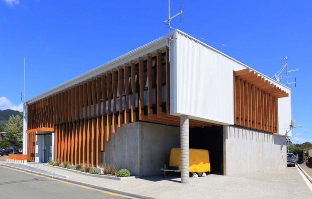 A contemporary two-storey building viewed from the street, with a strong geometric form, exposed concrete lower level, white corrugated upper cladding, and extensive vertical timber sun screens wrapping the glazed elevations. A covered parking bay is visible beneath the upper volume, with a paved forecourt and low landscaping. The image is an exterior architectural photograph under a clear blue sky.