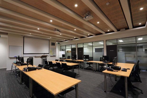 A modern training or conference room with multiple rectangular timber desks arranged in rows, black office chairs, desktop computers, a projection screen, whiteboard, and glass-partitioned rooms along the rear wall. The space has a dark carpeted floor, neutral walls, and a distinctive slatted timber ceiling with recessed lighting and exposed beams.