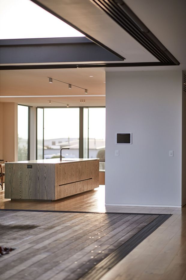 A bright contemporary open-plan kitchen viewed from an adjoining interior space. The kitchen features a large wood-grain island with integrated drawers and a slim dark benchtop, pale timber flooring, full-height glazing, minimalist track lighting, and a dramatic linear ceiling opening or skylight with dark framing. A blank wall with a small wall-mounted control screen separates the kitchen from the foreground.