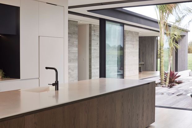 Contemporary kitchen with a large pale stone island benchtop, integrated sink and matte black tap, dark timber-look island cabinetry, and minimal white wall cabinetry. Full-height black-framed sliding glazing opens toward a timber outdoor deck and landscaped garden, creating a strong indoor-outdoor connection. The space is bright, clean-lined, and refined.