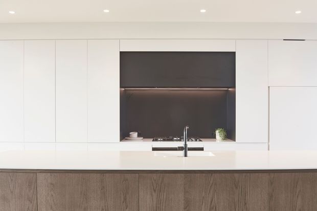 Minimalist contemporary kitchen with extensive matte white full-height cabinetry, a large dark feature splashback with integrated warm under-cabinet lighting, a pale stone island benchtop, and a timber-look island base. The composition is clean and streamlined, with recessed ceiling lights and integrated appliances.