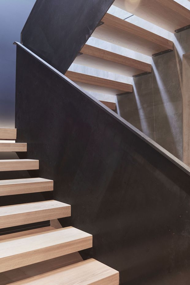 Close-up architectural view of a modern open-riser staircase. Light timber treads are cantilevered from a dark, solid-looking stair stringer or wall, with dark metal brackets and a slim metal edge detail. A pale stone or concrete wall forms the background, and the stair turns upward through the frame.