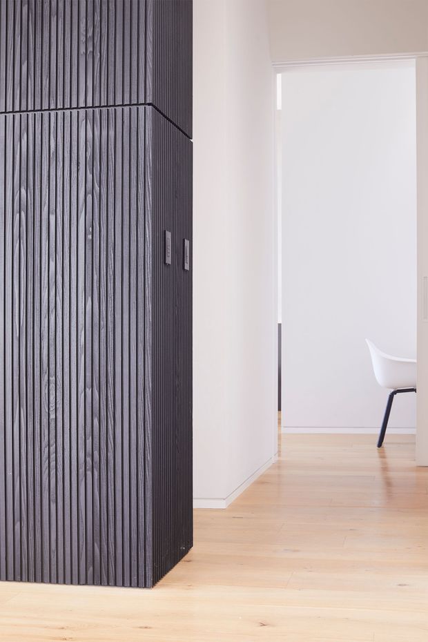 Minimalist interior view featuring a tall, dark vertically ribbed feature wall or built-in partition with integrated electrical controls. Pale timber flooring, white plaster walls, and a bright adjoining room with a white molded chair create a clean, understated composition. The image is focused on the textured architectural wall surface and hallway-like circulation space.