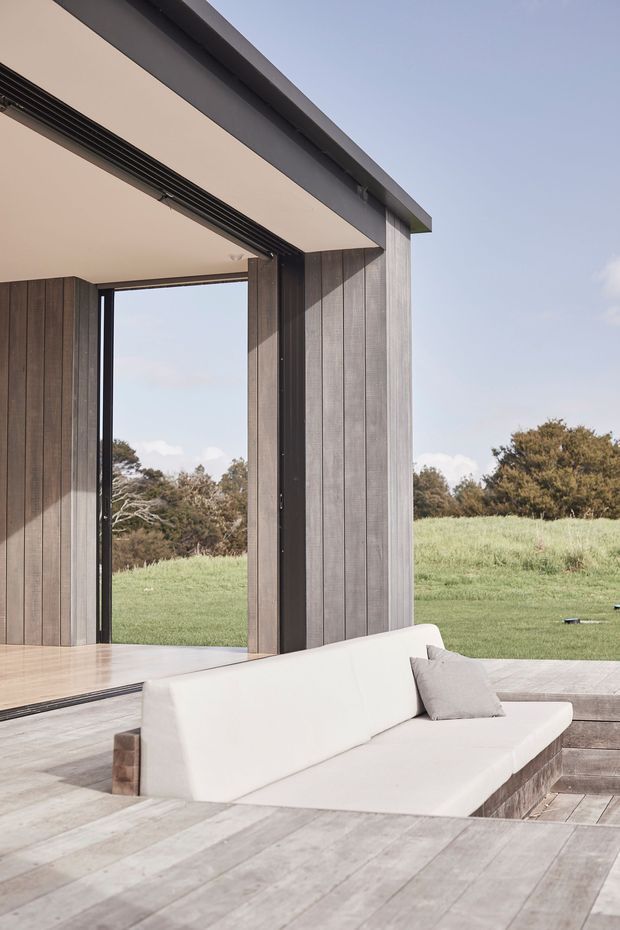 A contemporary open-air living area with a low white built-in sofa and cushion set into a broad timber deck. The space is framed by dark vertical timber wall cladding, slim black-framed full-height openings, and a flat projecting roof with a concealed linear ventilation grille. A grassy rural landscape is visible beyond the open glazing, creating a strong indoor-outdoor connection.