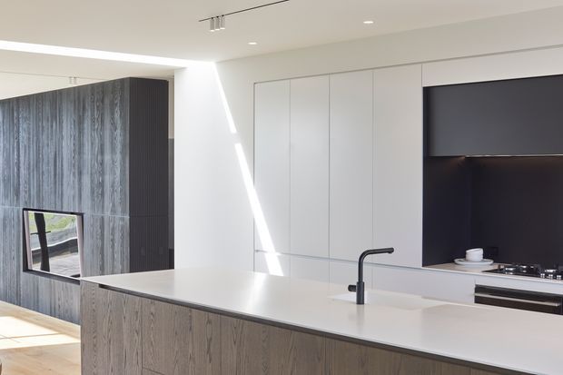 A bright, minimalist contemporary kitchen with a large island featuring a pale stone benchtop and vertically grained timber cabinetry. Full-height white cabinetry lines the rear wall, with a dark integrated cooking and splashback zone. A tall dark timber-clad volume with an inset television or reflective appliance is visible on the left. Recessed and slim track lighting, clean lines, and strong natural light create a crisp, refined interior.