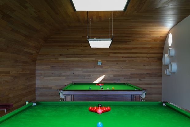 A dedicated billiards room with two green-felt pool tables aligned end-to-end, surrounded by warm horizontal timber wall and ceiling lining. Suspended rectangular billiard lights illuminate the tables, while a curved white wall with circular accents forms one side of the room. The space has a dark, warm, recreational atmosphere.